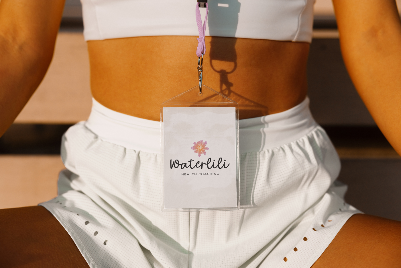 Close-up of a lanyard badge for Waterlili Health Coaching resting against the waist of a woman dressed in white athletic wear. The badge features a colorful lotus-style logo and brand name, with the person seated cross-legged in a relaxed pose.
